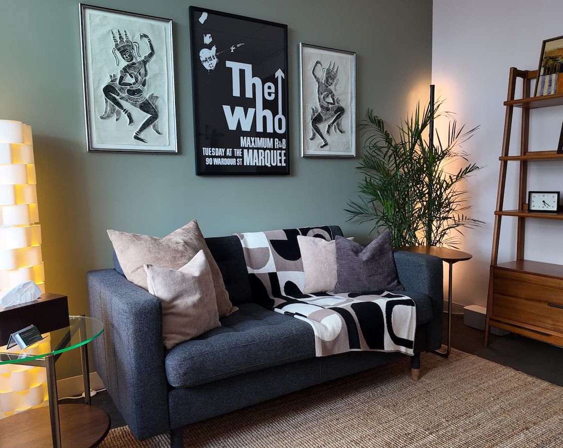 Client seating area with gray sofa, wooden bookshelf unit, palm plant, geometric throw blanket, and framed artwork on the wall.