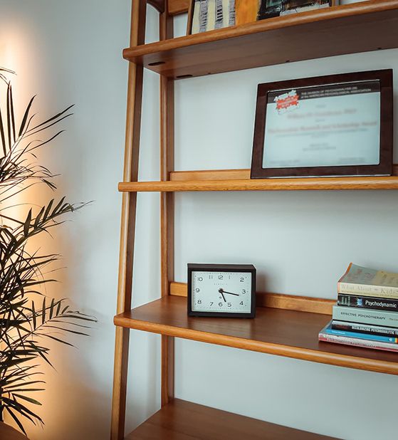 Close-up view of wooden bookshelf with psychology and therapy books, a desk clock, and framed certificate, with a large palm plant in the corner