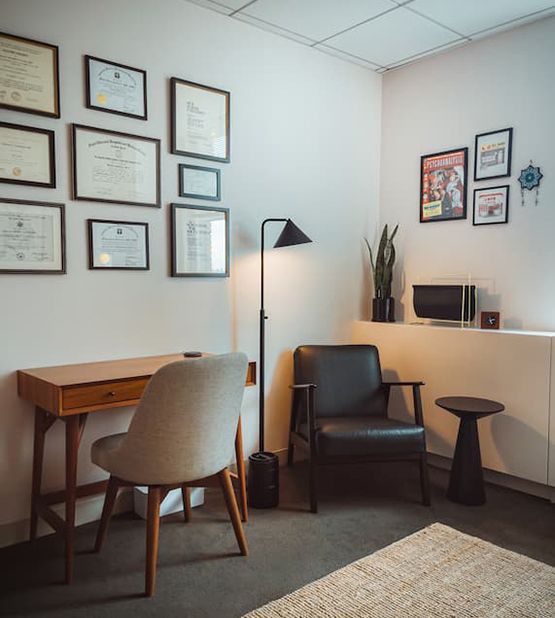 Therapist office interior showing a wooden desk with modern chair, black leather therapist chair in the corner, floor lamp, and wall displaying framed professional certificates and diplomas